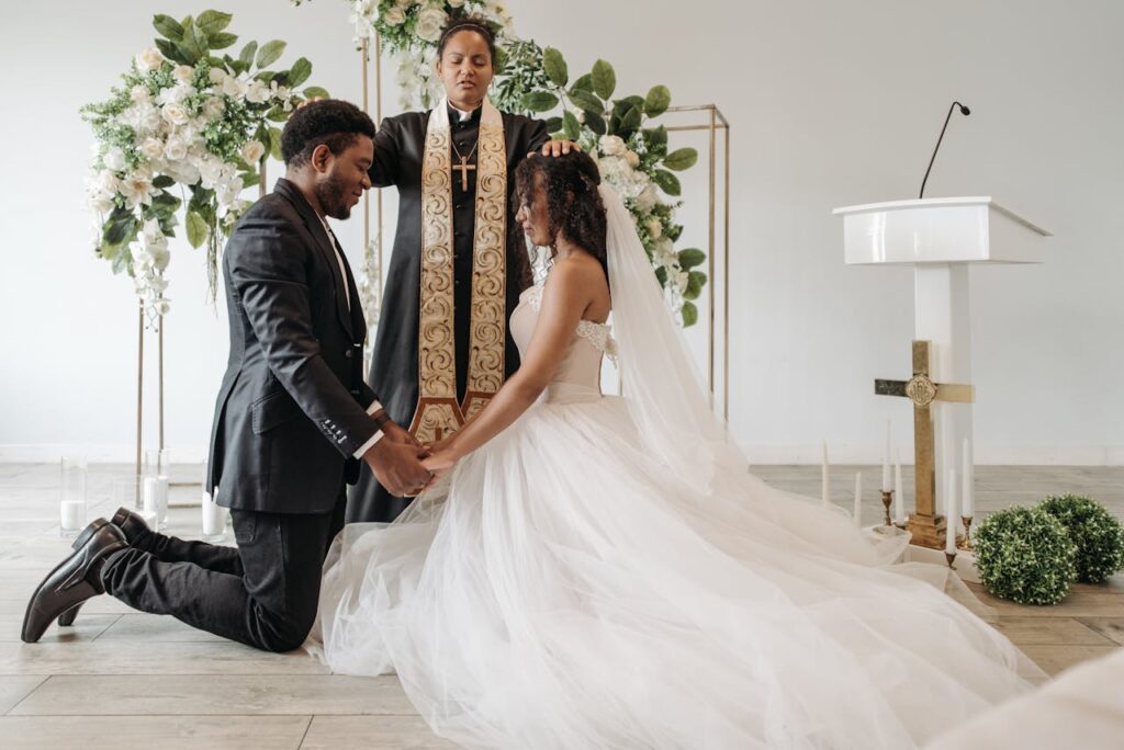 Beautiful indoor wedding ceremony with a bride and groom kneeling in prayer before a pastor.