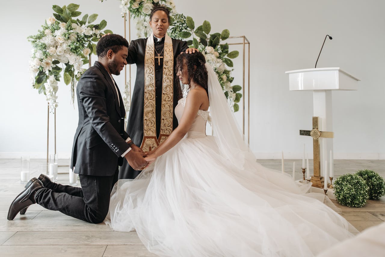 Beautiful indoor wedding ceremony with a bride and groom kneeling in prayer before a pastor.